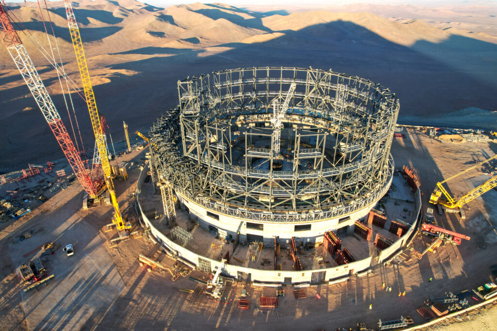 Denna bild togs i juni 2023 och visar en webbkamerabild av konstruktionsplatsen för ESO:s ELT i Chiles Atacamaöken. Stålskelettet kommer att vara den bärande strukturen för kupolen som ska vara hem till 39-meters teleskopet. Byggarbetare i bildens nedre kant ger skala åt den enorma konstruktionen, Bortom kupolen ser vi skuggan av Cerro Armazones, berget ELT byggs på. Källa: ESO